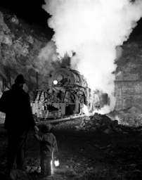 This photograph shows C.E. “Jack” Hash, a member of the Norfolk & Western Advertising department, holding hands with his young son while watching a class Y5 locomotive emerge from the Montgomery Tunnel in Christiansburg. It is one of O. Winston Link’s most popular images, demonstrating Link’s skills as a night photographer and capturing the ageless human fascination with trains. 	