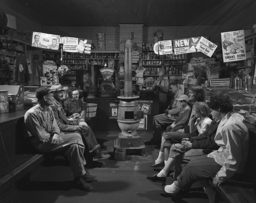 A group of white Virginians, two of them children, most of them dressed casually or in work clothes, sit across from one another on two benches in a train station around a woodburning stove. A man in the back appears to be taking a grocery order.  