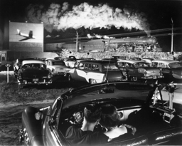 A white couple cuddle in a convertible at a packed drive-in movie, while a steam train roars by in the background. At the moment the picture was taken, a jet appeared on screen, so the image captures cars, steam trains, and a jet in one photograph. 