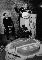 This photograph, taken for the Richmond Times-Dispatch, shows a Republican Party reception held at the John Marshall Hotel in February of 1977. Then California governor Ronald Reagan sits next to a podium, where a speaker is presenting him with a plaque. A large cake in the shape of an elephant decorated with three stars, a Republican Party symbol, sits in the foreground of the image. 