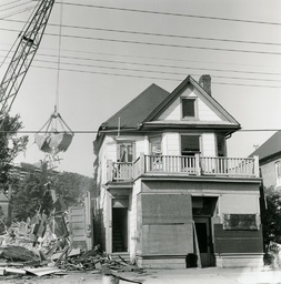 <p>A crane demolishes a building behind a boarded-up house in this photograph of East Ghent, Norfolk, part of the collection of the Norfolk Redevelopment and Housing Authority (NRHA)."